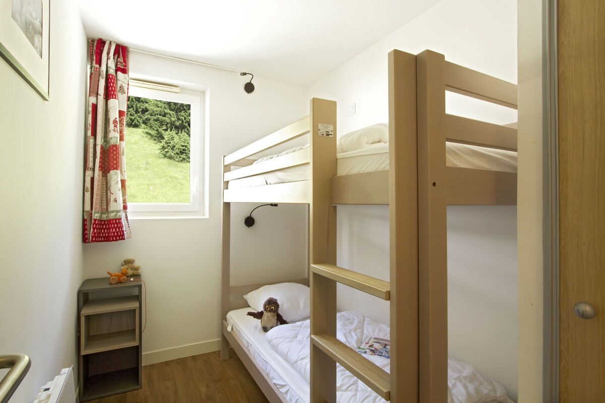 Una habitación pequeña y luminosa con una cama litera, una mesita llena de juguetes y una ventana que da a zonas verdes.