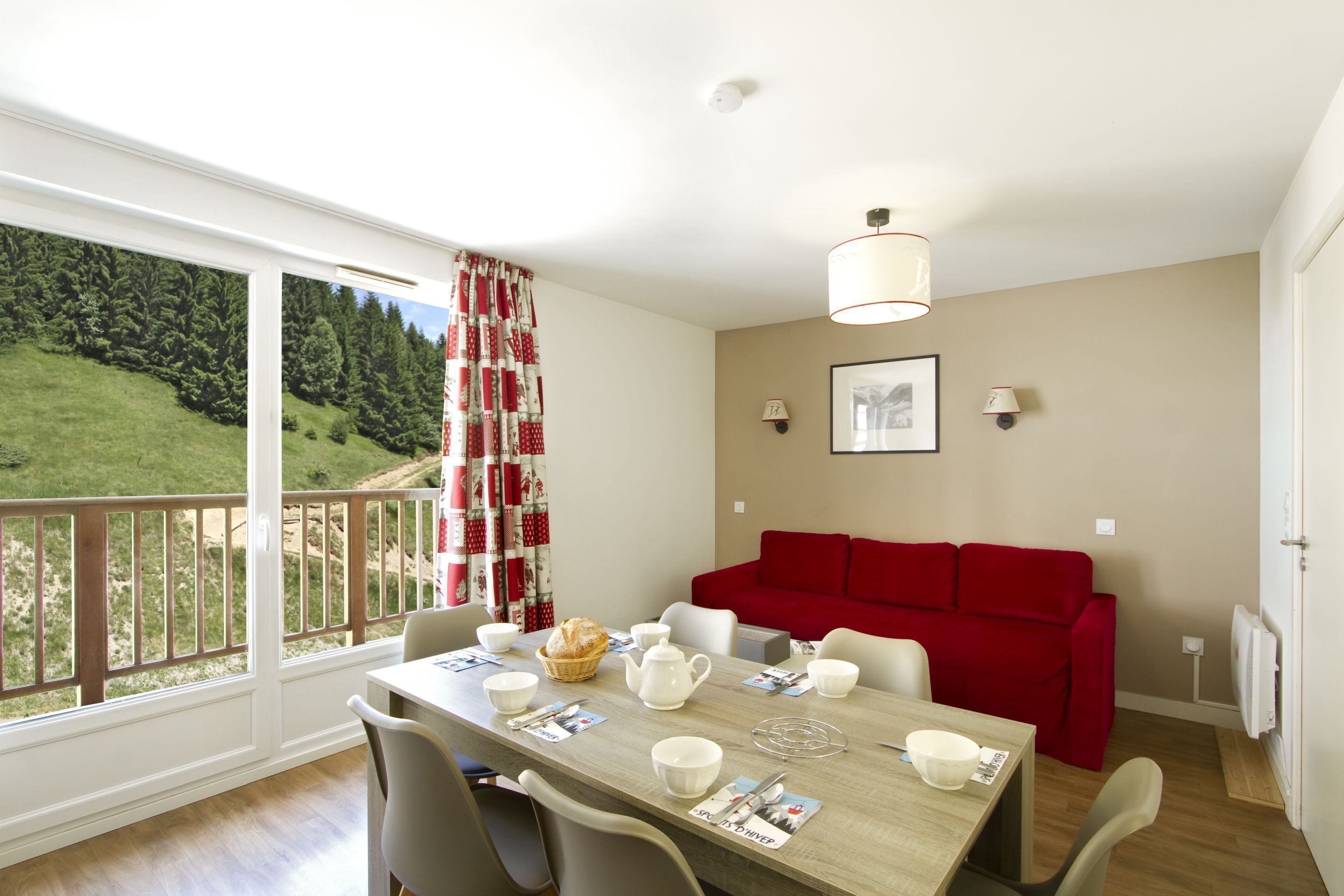 Un coin repas avec une table dressée pour quatre personnes, un canapé rouge et une vue sur un paysage verdoyant, visible à travers de grandes fenêtres.