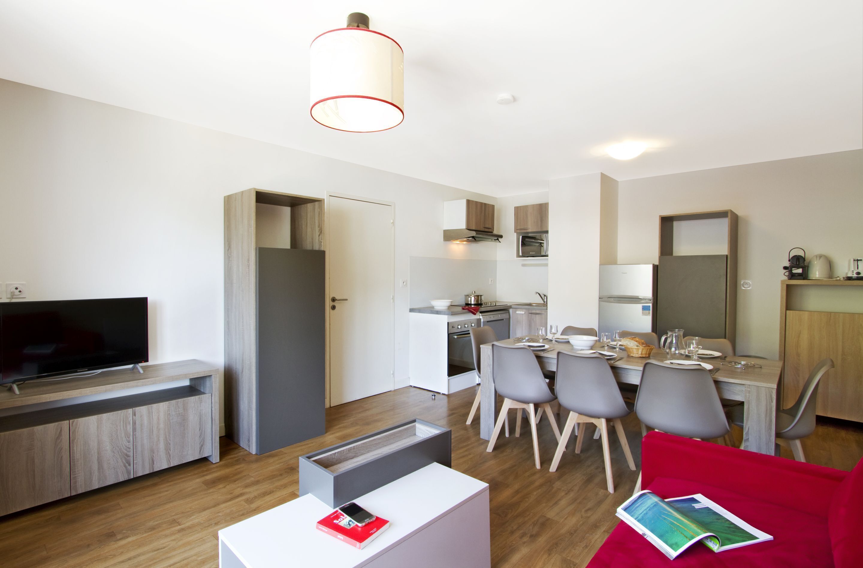 A modern living room with a flat-screen TV, a dining area with a table set for four, and a kitchen with a stove and oven.