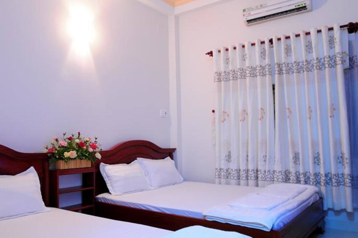 A hotel room with two beds, a nightstand with a flower arrangement, and a window with patterned curtains.