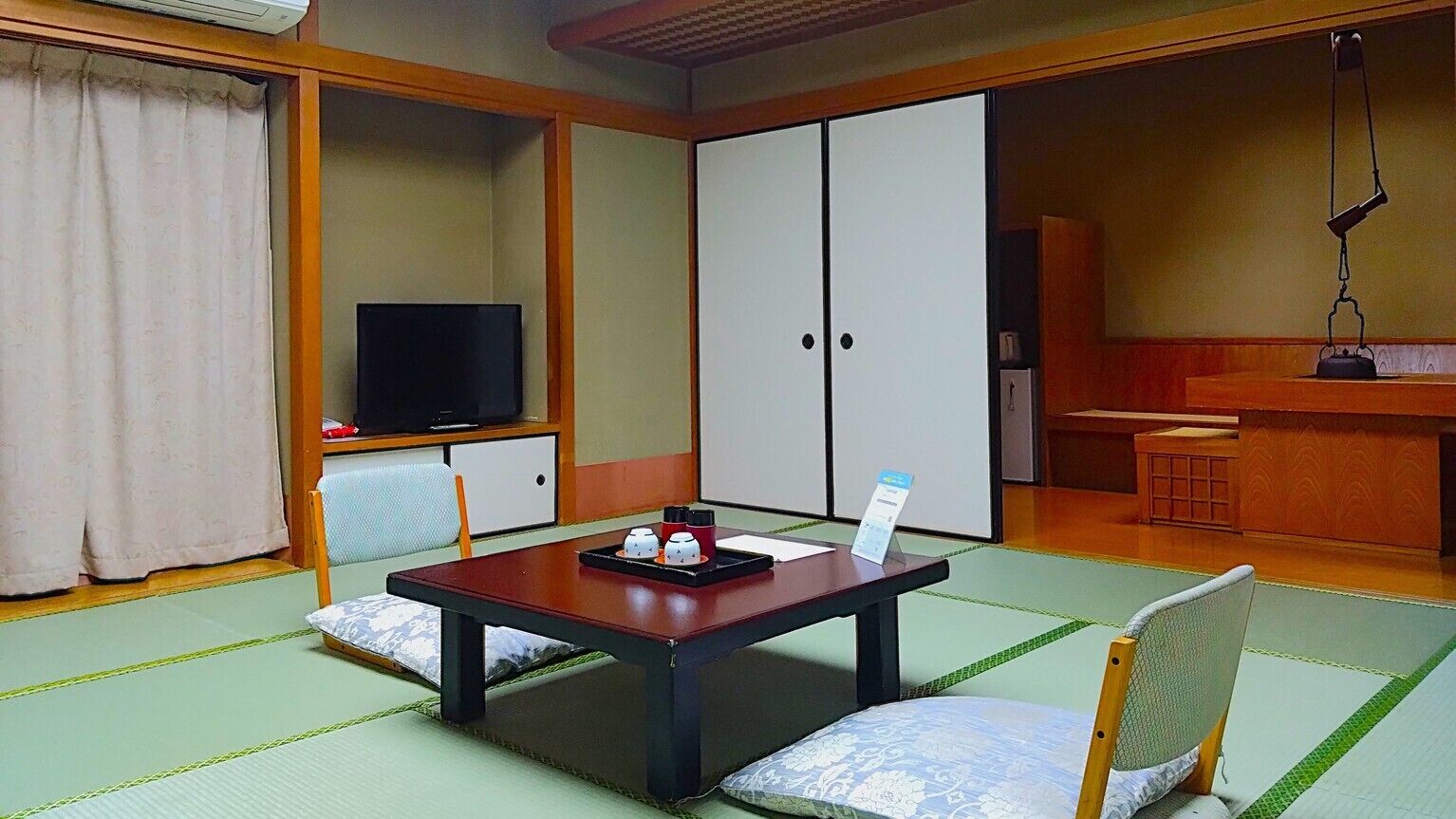 Um quarto japonês tradicional com piso de tatame, uma mesa baixa com bandeja, televisão, portas de correr e uma pequena cozinha.