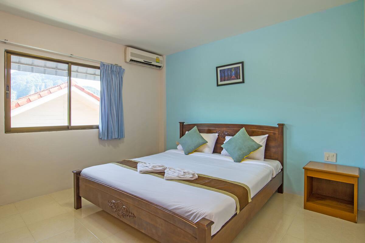 A neatly made bed with pillows and a set of towels, a wooden nightstand, a window with curtains, and an air conditioning unit.