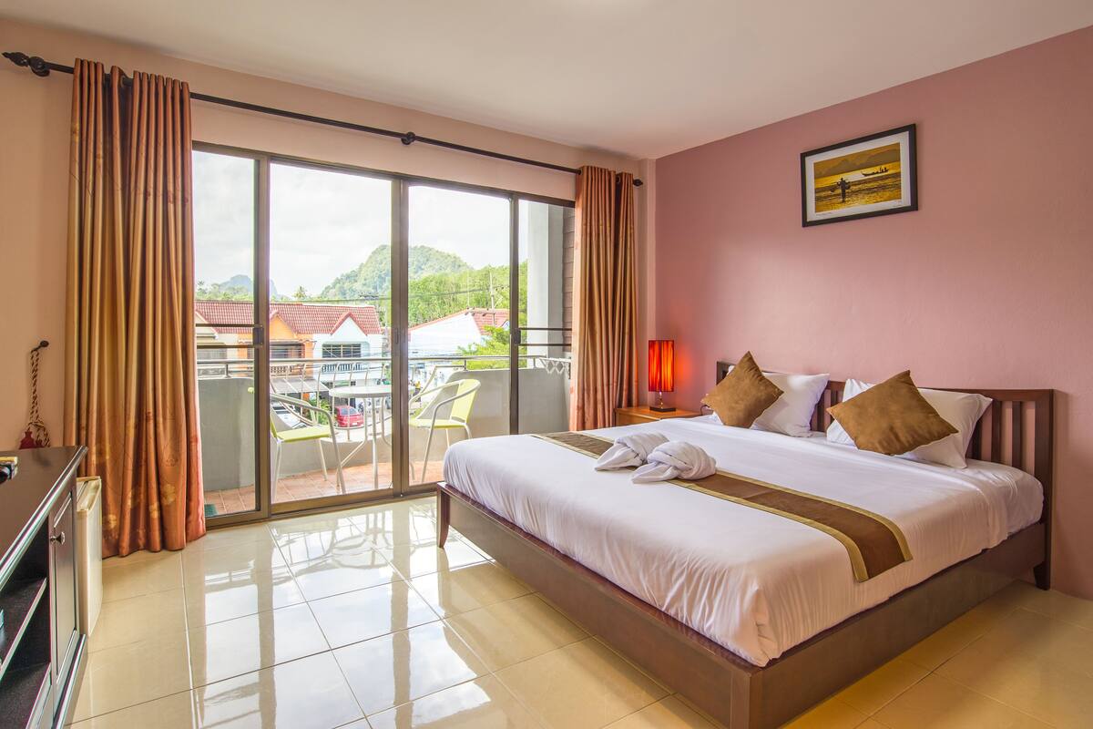 A hotel room with a large bed, a balcony with a table and chairs, and a view of the surrounding area.