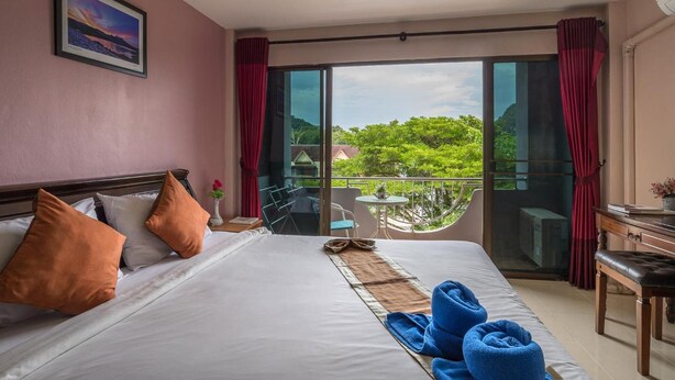 A hotel room with a bed, pillows, a view of greenery, a balcony, and a table with a chair. - Mukanda Residence (Krabi)