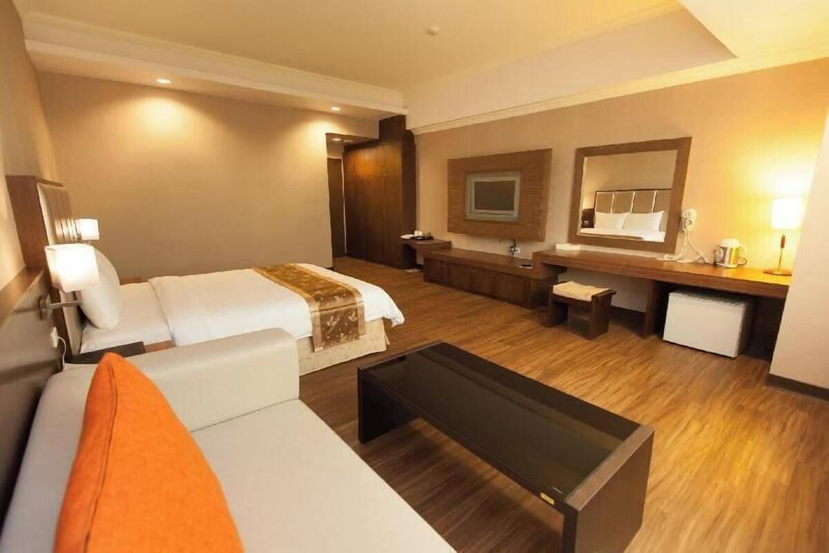 Habitación de hotel con cama, escritorio, espejo, una mesita pequeña y baño con lavabo y espejo.