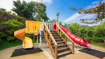 Children can play on the slide and climbing structure amidst lush greenery and trees.