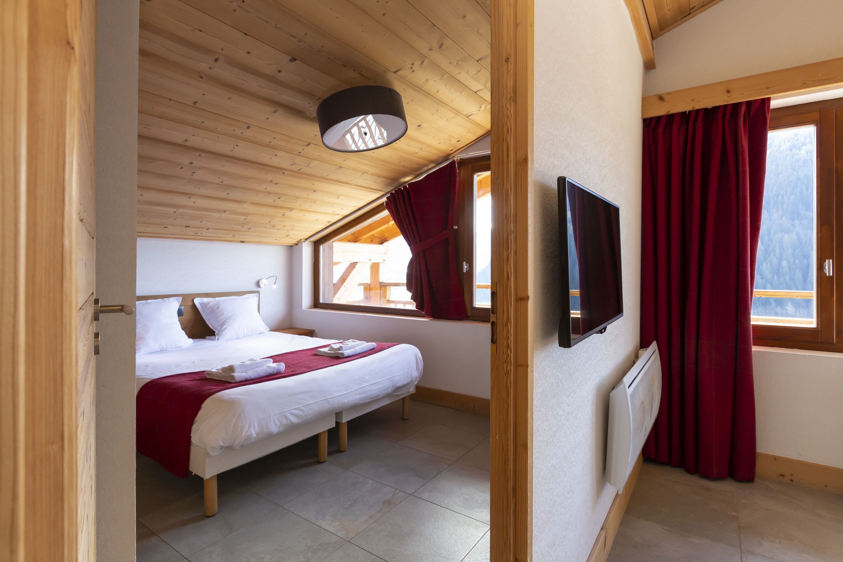 A bedroom with a bed, a TV, a window with curtains, and a wooden ceiling.