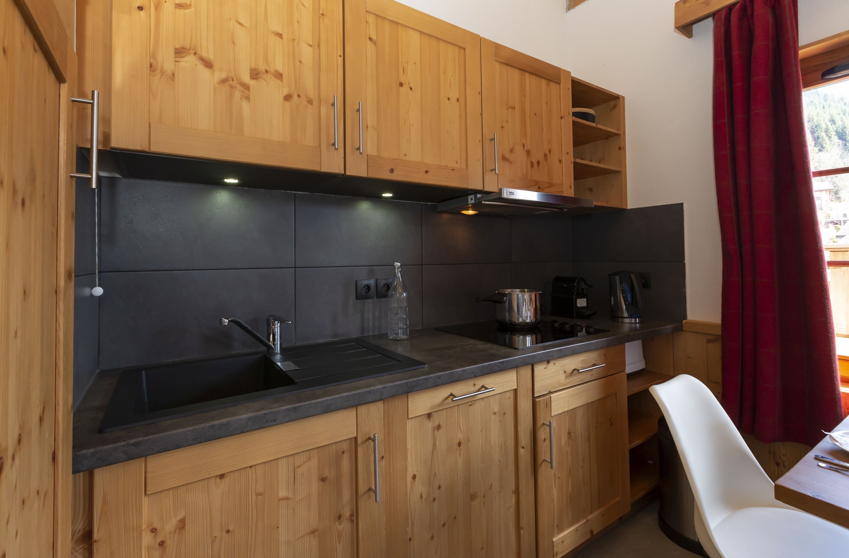 Une cuisine avec des meubles en bois, un plan de travail noir, un évier, une cuisinière et une fenêtre ornée de rideaux rouges.