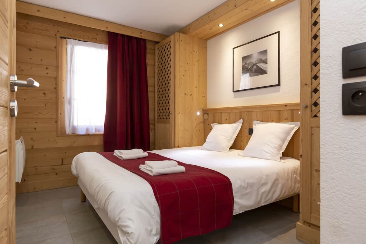 Een kamer met houten lambrisering, een bed, een raam met gordijnen, een foto in een lijst aan de muur en een thermostaat.