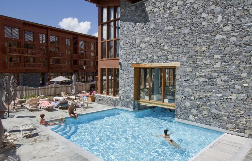 Un espace piscine avec des chaises longues et des parasols, un bâtiment en pierre doté de grandes fenêtres et une structure en bois avec des balcons.