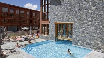 Un espace piscine avec des chaises longues et des parasols, un bâtiment en pierre doté de grandes fenêtres et une structure en bois avec des balcons.