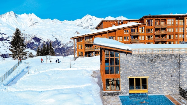 Un hotel con piscina, pista de esquí y montañas nevadas al fondo.