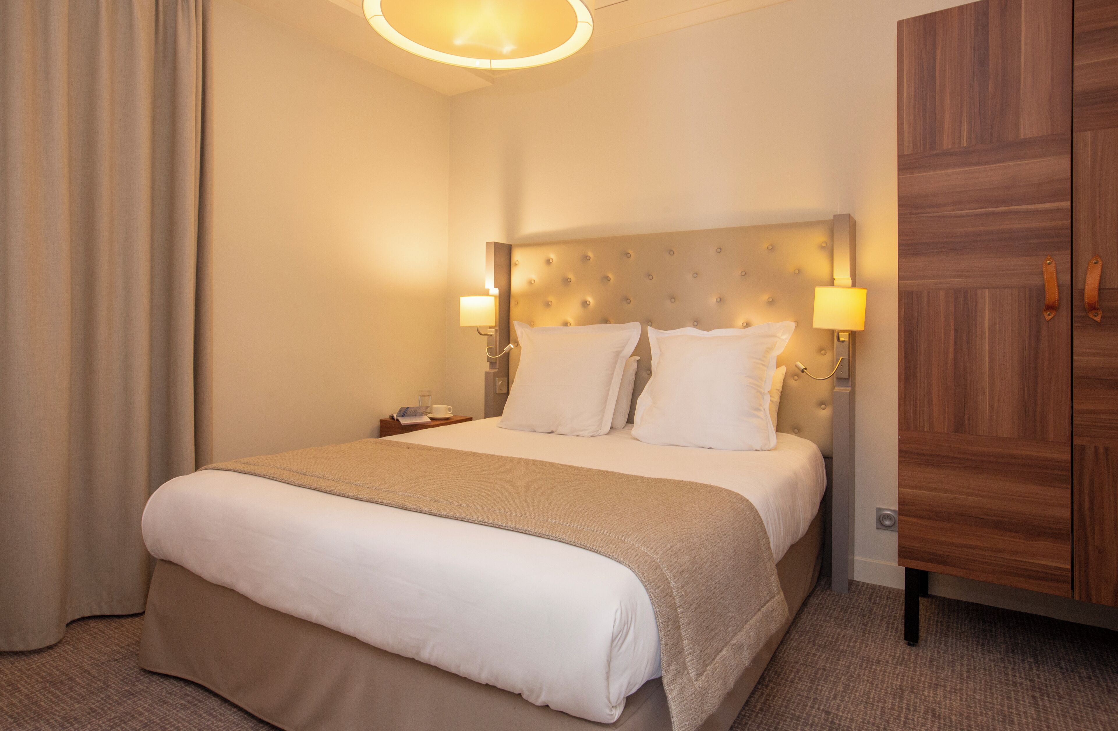 A hotel room with a large bed, two bedside lamps, a wooden wardrobe, and a neutral color scheme.