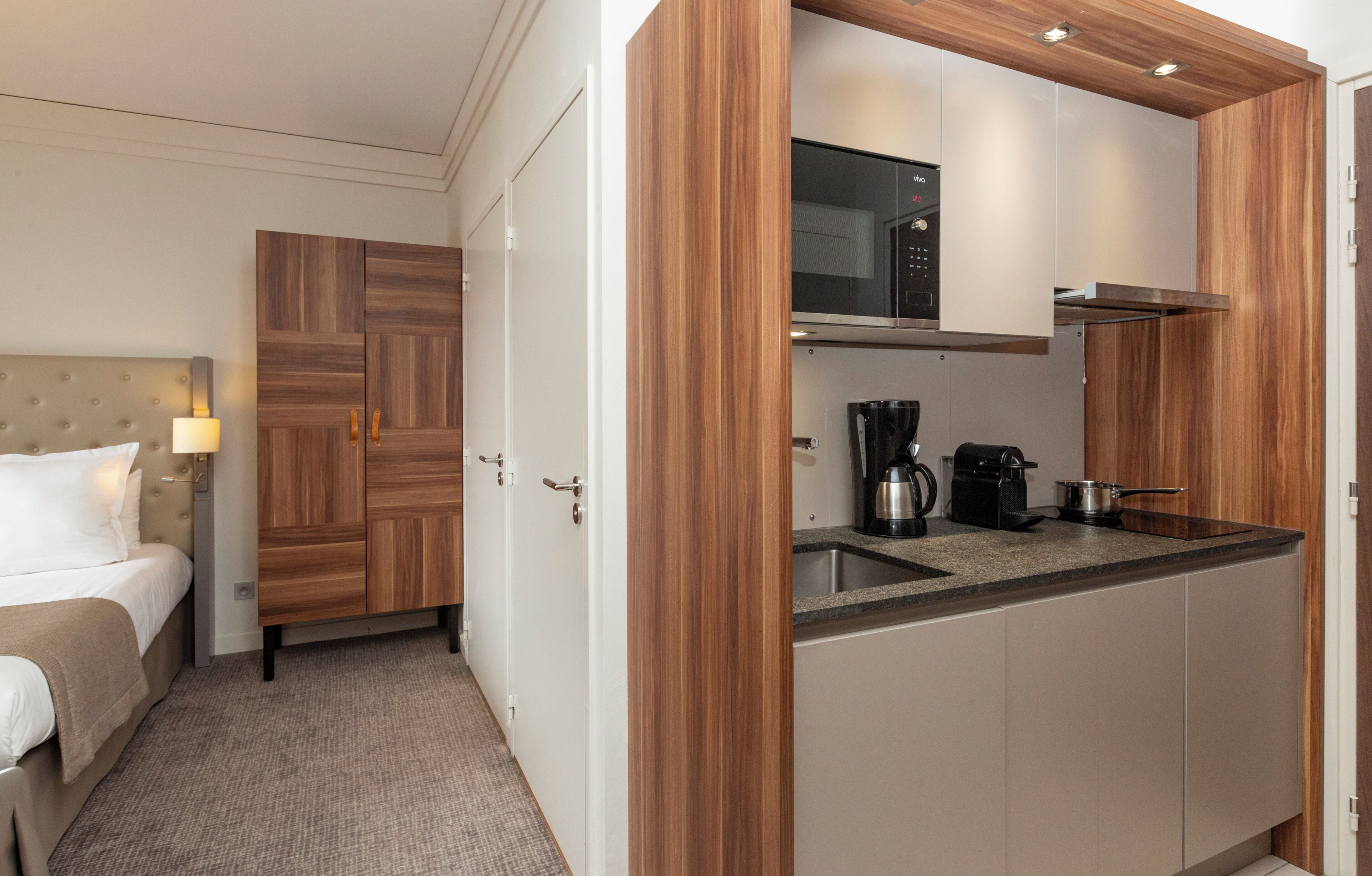 A hotel room with a bed, a wooden wardrobe, a kitchenette with a microwave, a coffee maker, and a sink.
