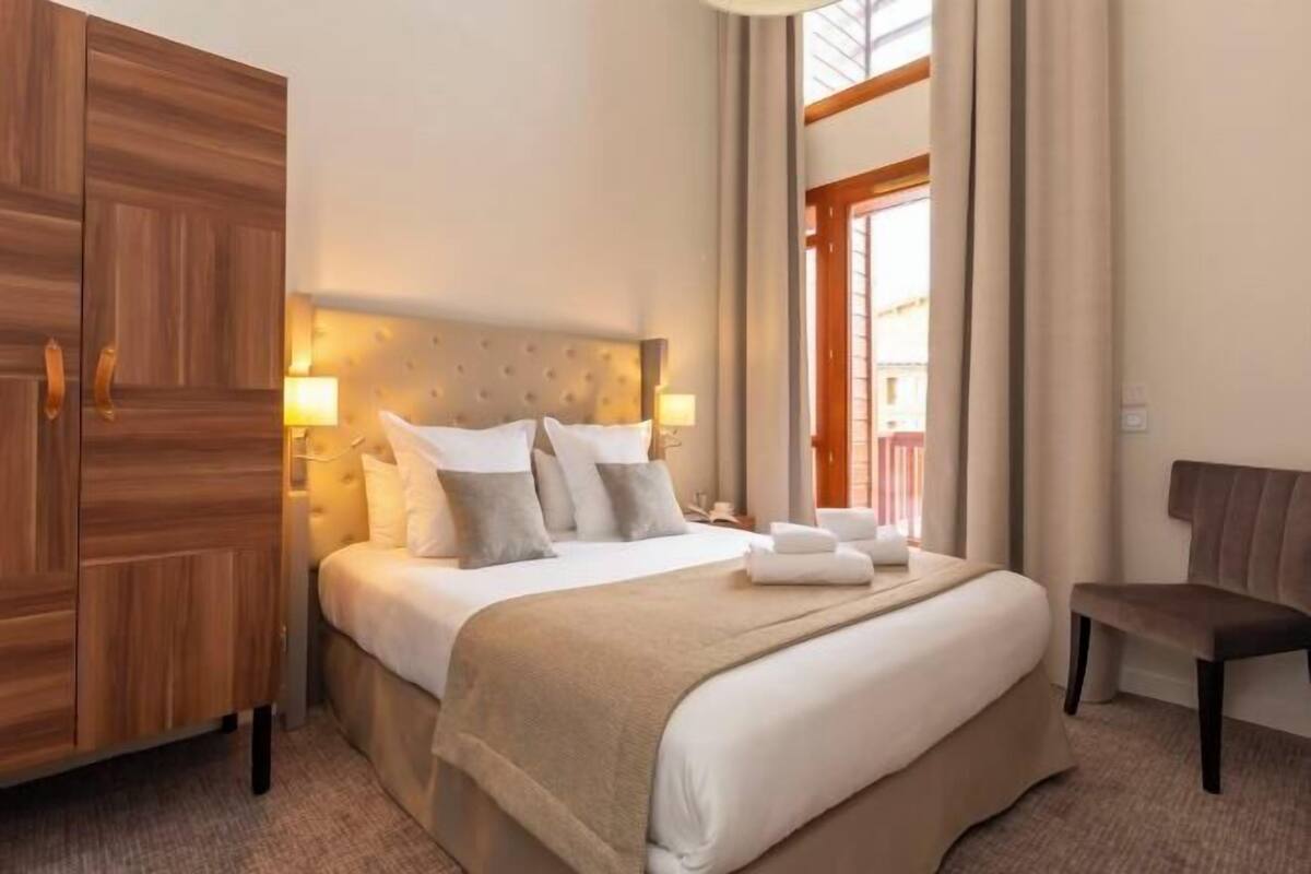 Een hotelkamer met een groot bed, bedlampjes, een houten garderobe, een bankje en een raam met gordijnen.