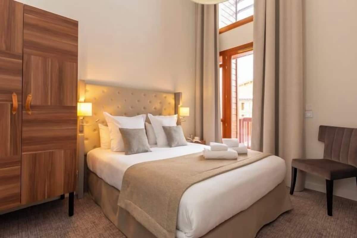 A hotel room with a large bed, bedside lamps, a wooden wardrobe, a bench, and a window with curtains.