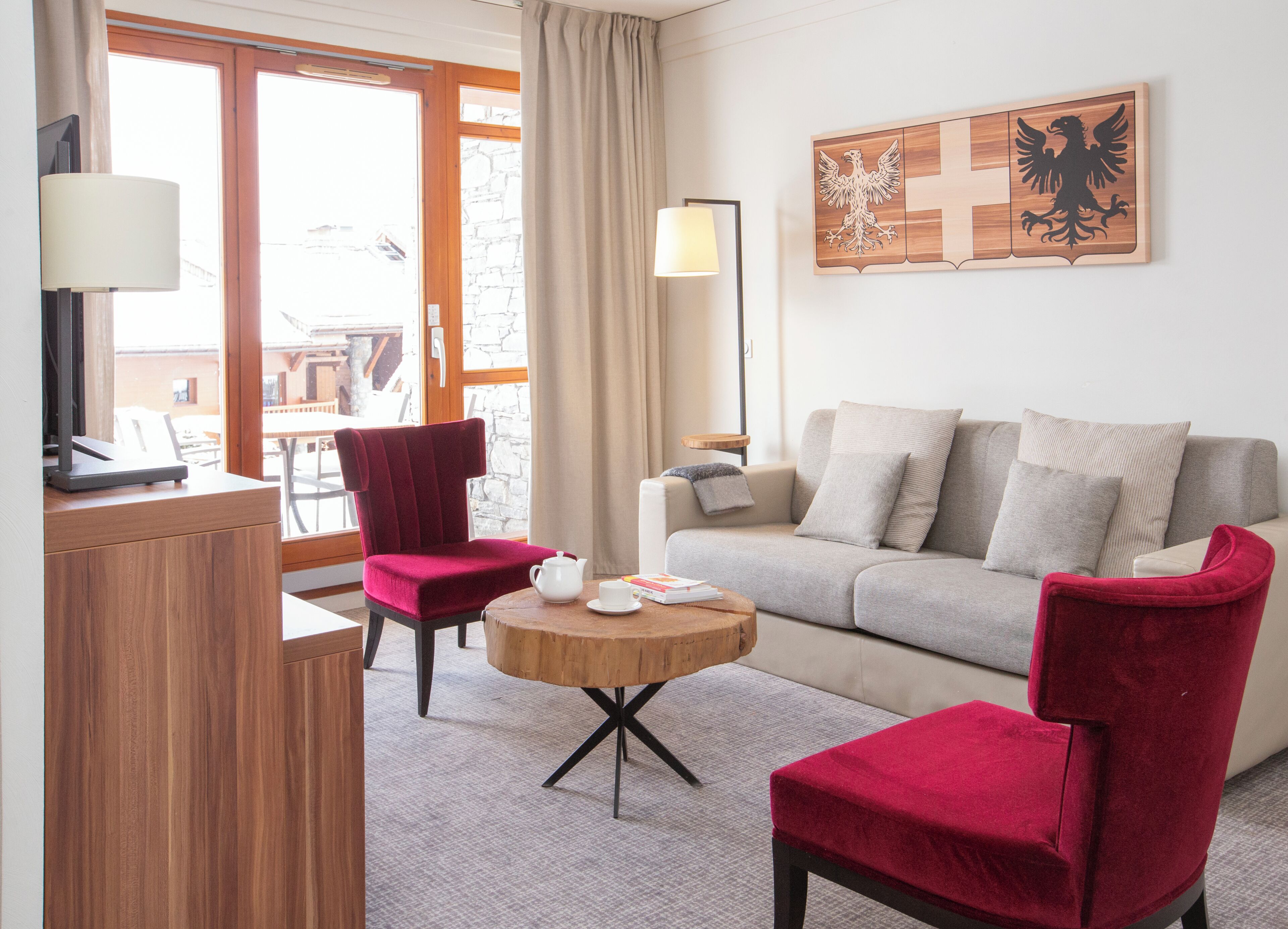 A hotel room with a sofa, two red chairs, a small wooden table with a teapot, a lamp, and a wall-mounted artwork.