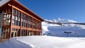 Un edificio moderno con amplios ventanales y fachada de madera, con imponente fondo de montañas nevadas.