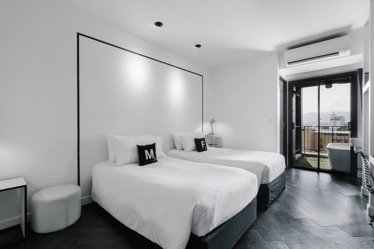 A modern hotel room with two beds, a small round ottoman, a nightstand, a desk, and a balcony with a view of the city.