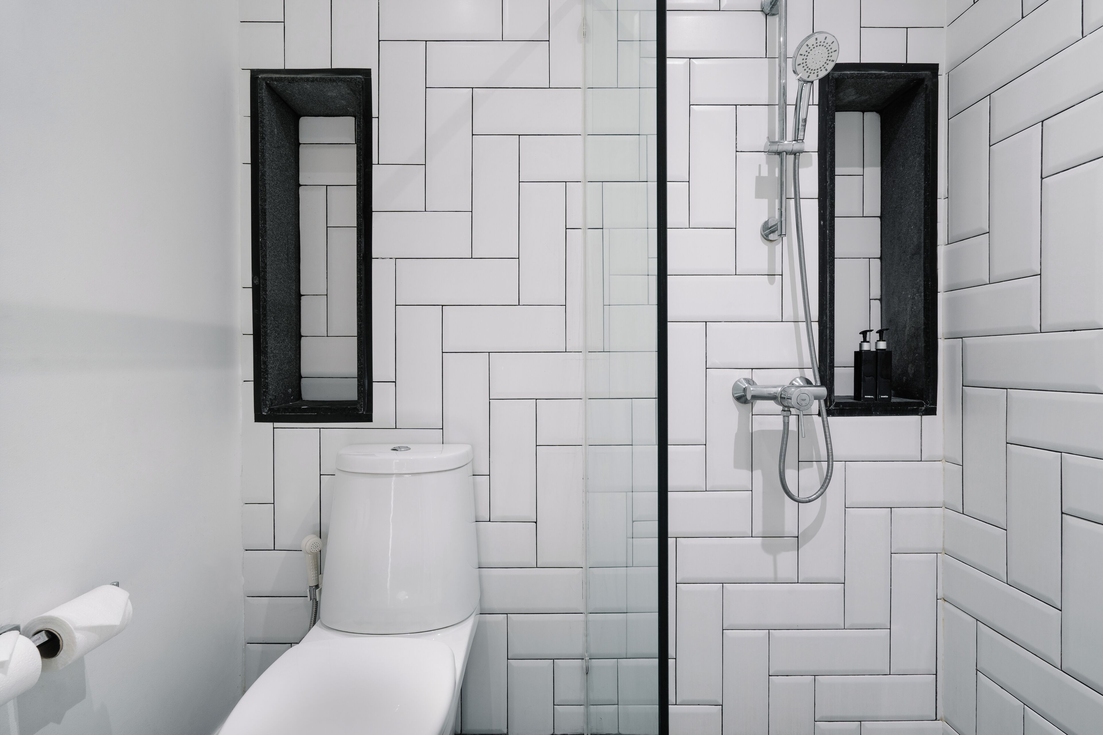 Une salle de bain moderne avec des carreaux blancs disposés en motif chevrons, des toilettes, une douche avec un pommeau de douche amovible et deux miroirs à cadre noir.