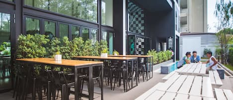 An outdoor seating area with tables and chairs, surrounded by greenery and large windows.