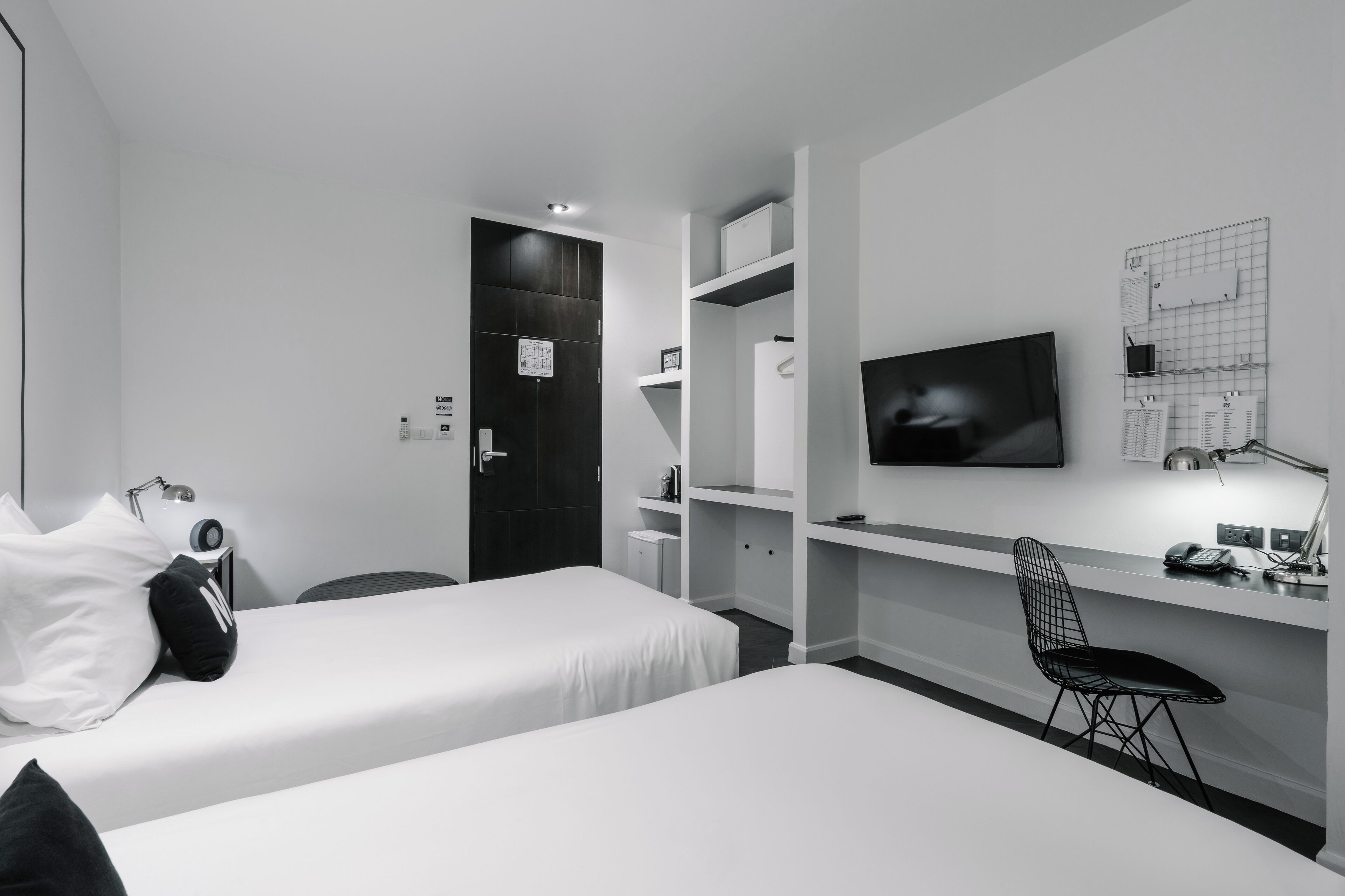 A modern hotel room with two single beds, a desk with a chair, a TV, and built-in shelving.