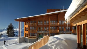 A multi-story wooden building with balconies, a snow-covered landscape, and mountains in the background. - Résidence Odalys Edenarc - Arc 1800 (Bourg-Saint-Maurice)