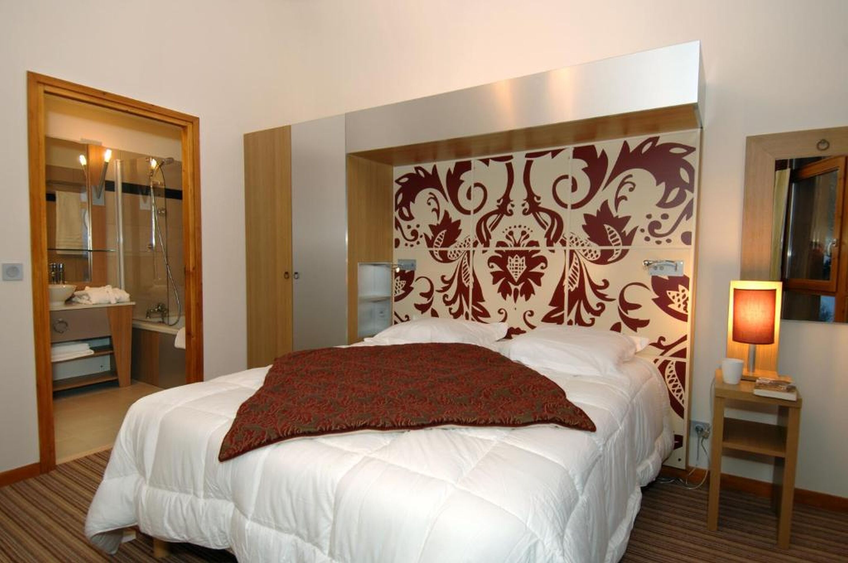 A hotel room with a large bed, a mirror, a bedside table with a lamp, and a bathroom visible through an open door.