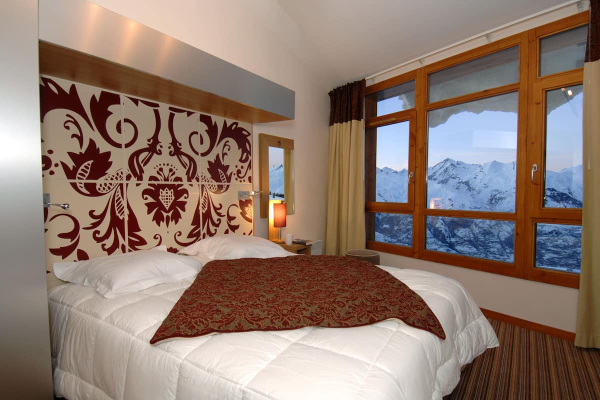 A hotel room with a large bed, a decorative headboard, a nightstand with a lamp, and a window with a mountain view.