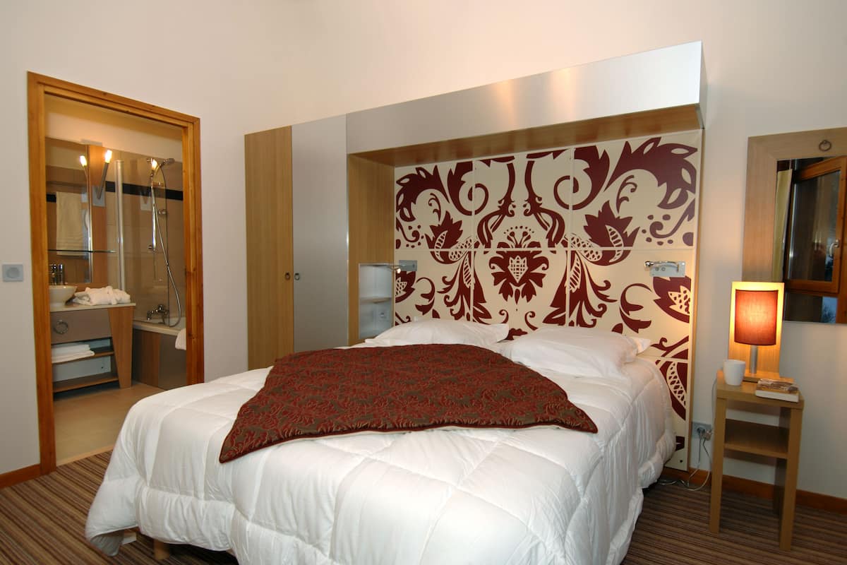 A hotel room with a large bed, a wooden headboard, a bedside table with a lamp, and a bathroom visible through an open door.