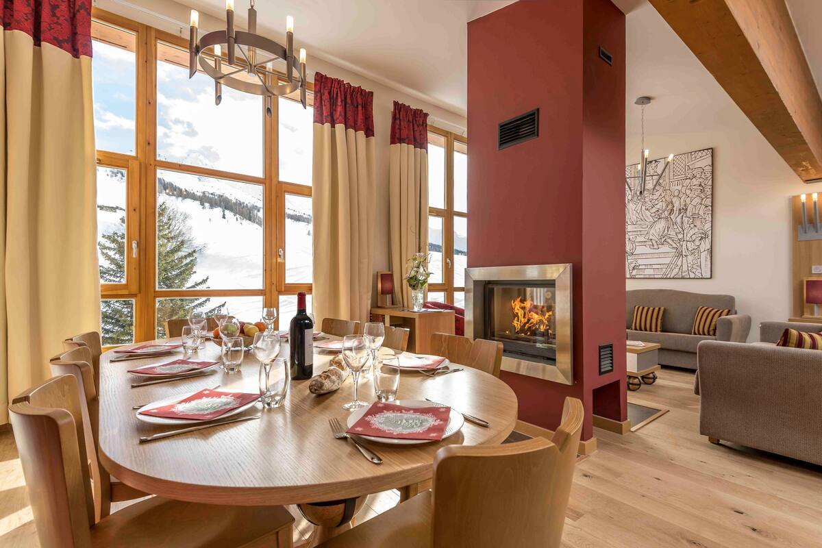 A dining area with a wooden table set for six, a fireplace, and a view of a snowy landscape through large windows.