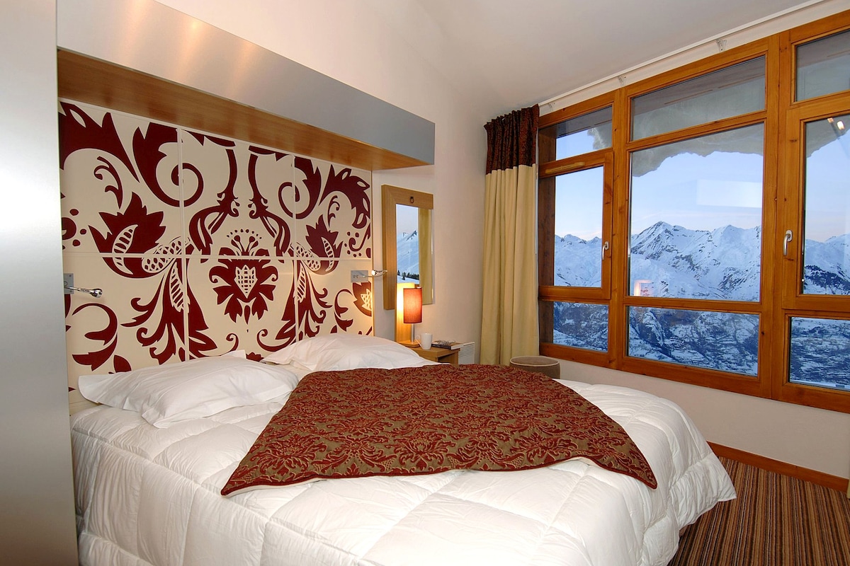 A hotel room with a large bed, a decorative headboard, a nightstand, a lamp, and a window with a mountain view.