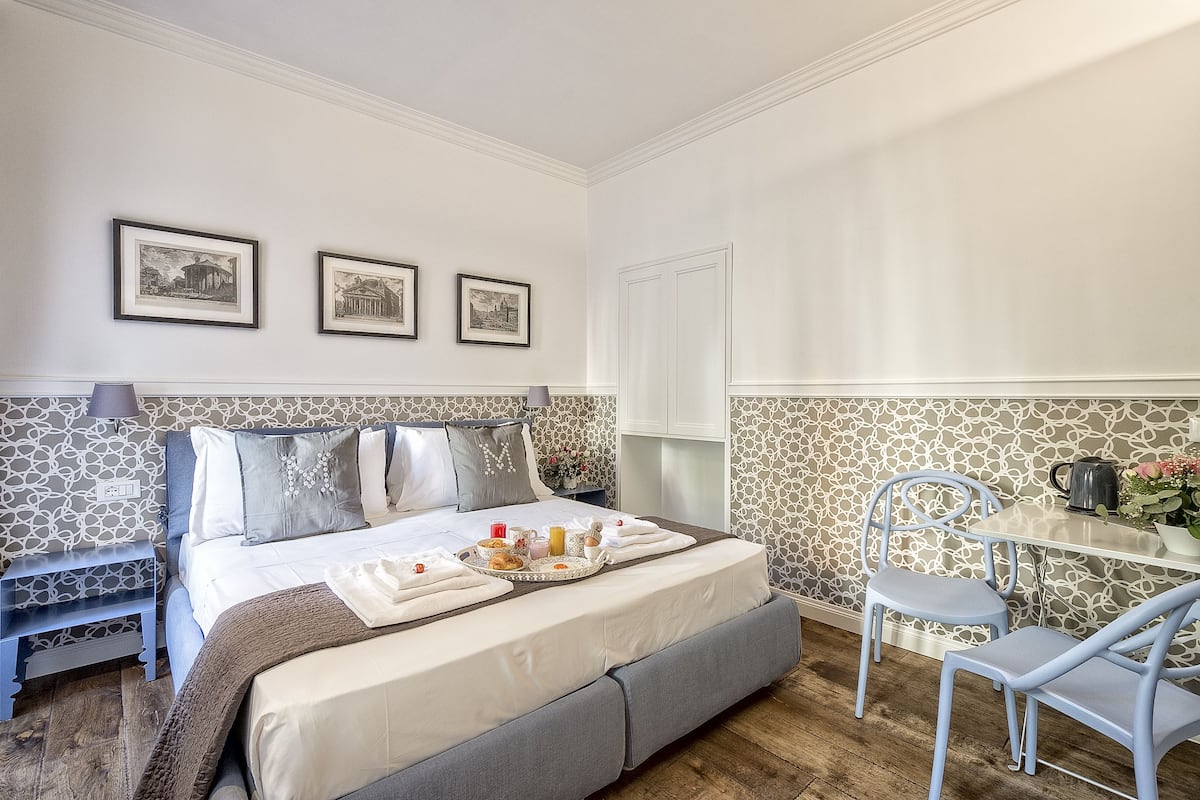 A neatly arranged bedroom with a bed, bedside table, and a small table with a kettle and flowers.