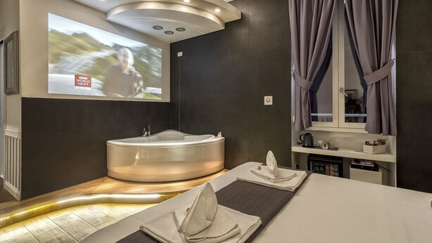 A modern bathroom with a large bathtub, a vanity with a mirror, and a projection screen displaying a news program. - Magenta Collection Sant'Anna (Rome)