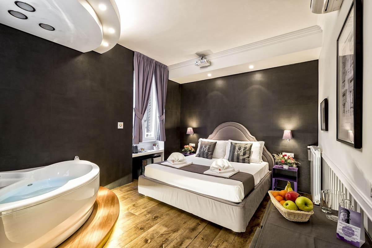 A modern hotel room with a large bed, a bathtub, a small desk, and a fruit basket.