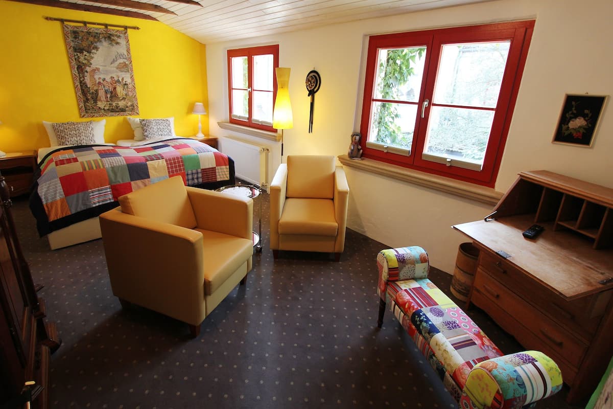 A room with a bed, two chairs, a wooden dresser, a lamp, and a window with red frames.