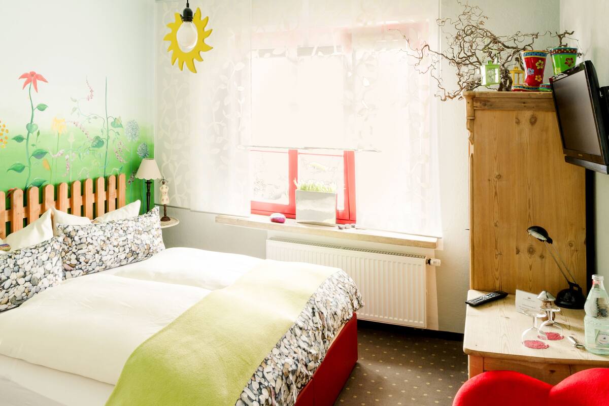 A bedroom with a bed, a desk, a TV, and a window with a view of greenery.