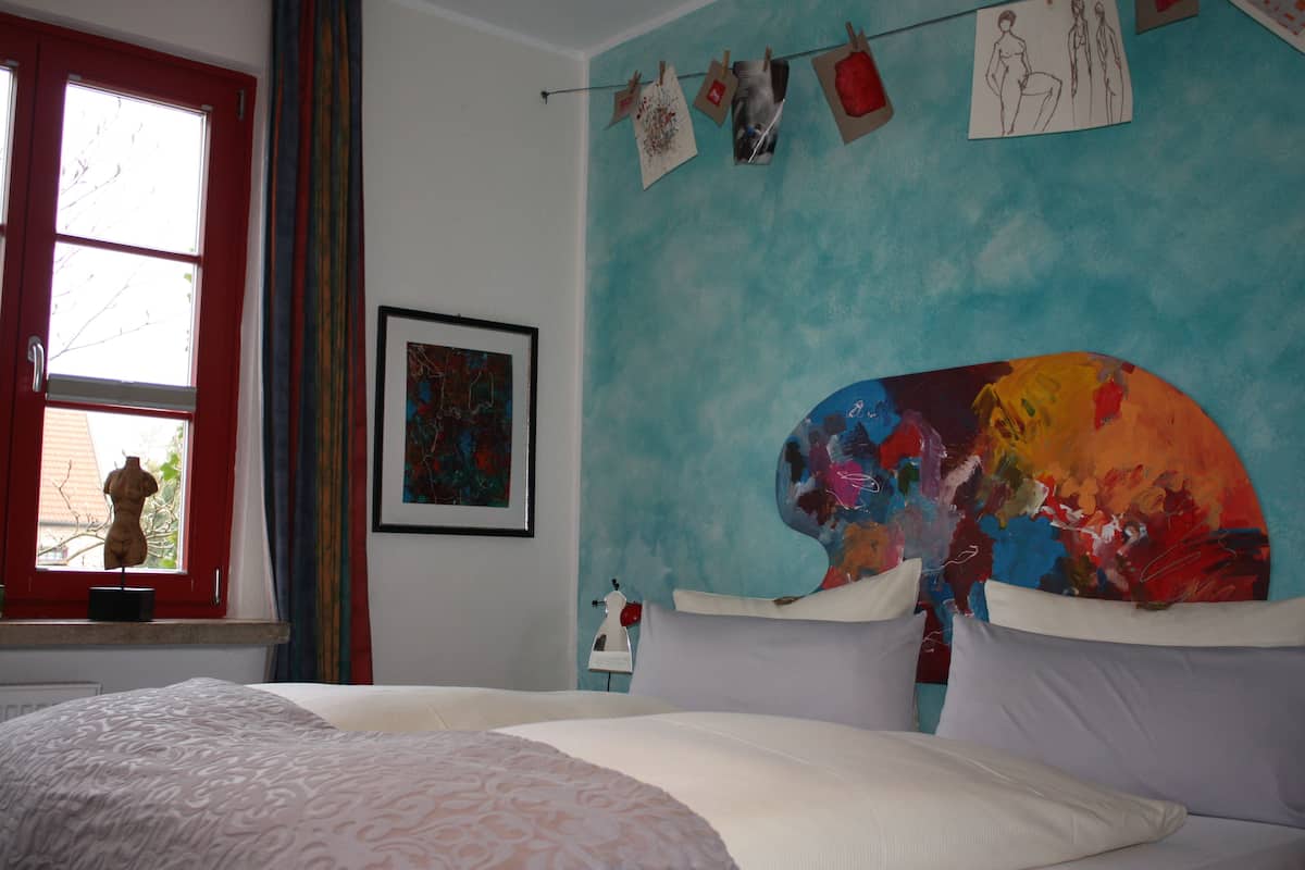 A bedroom with a bed, a window with curtains, a wall with a painting, and a mannequin on a shelf.