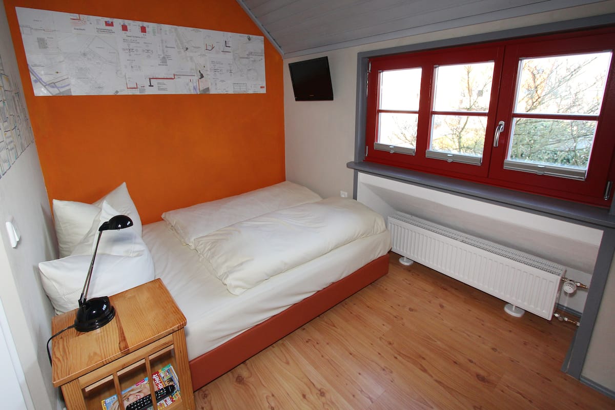 A bedroom with a single bed, a nightstand with a lamp, a window with red frames, and a radiator.