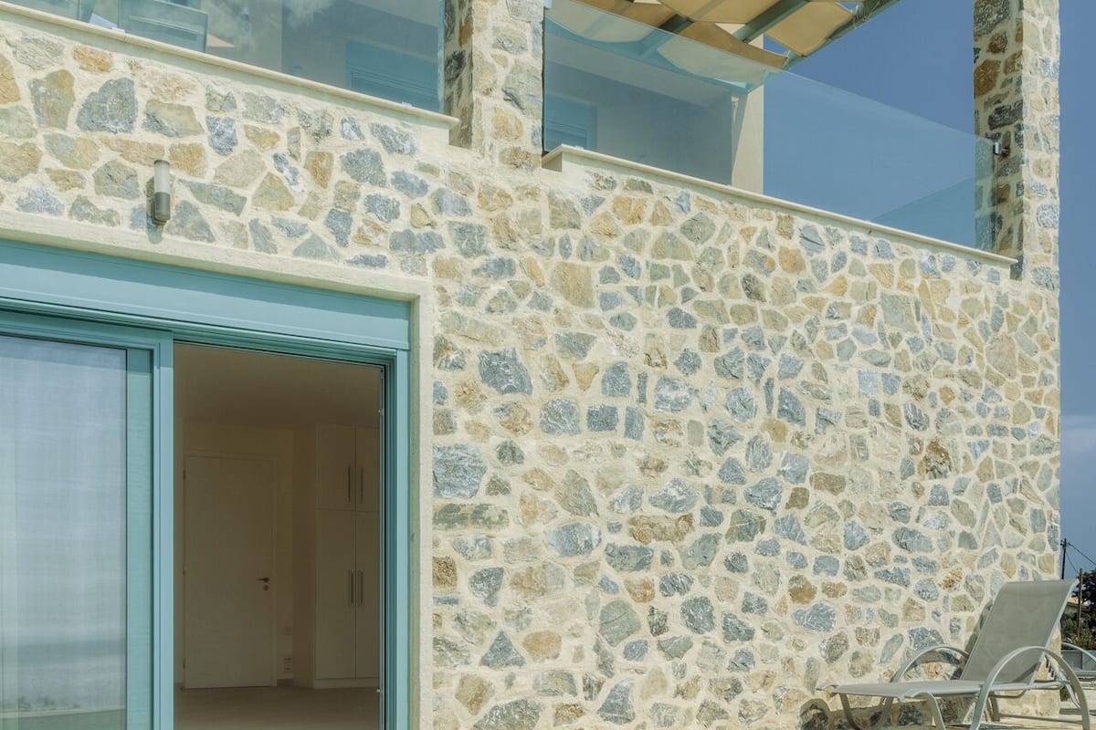 Una casa moderna con fachada de piedra, un portón de cristal que da a una habitación y una zona de piscina con una tumbona.