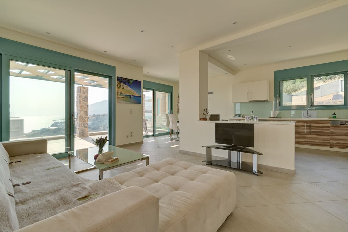 Una sala de estar moderna con un sofá, una mesa de centro y un televisor. Hay una cocina con un fregadero y armarios. La habitación tiene ventanas amplias con vista al mar.