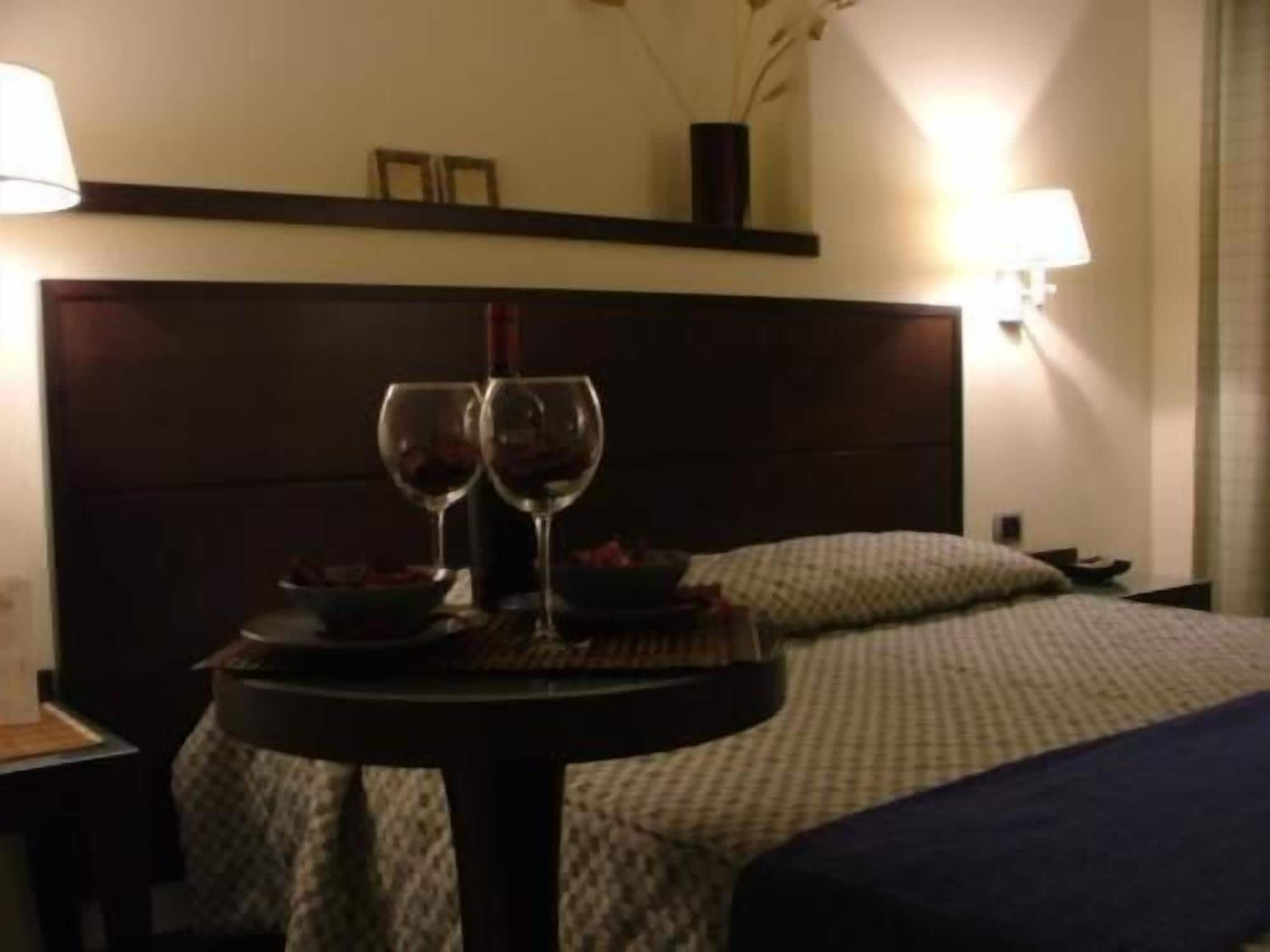 A hotel room with a bed, a bedside table with two wine glasses and a bowl, and a wall-mounted lamp.