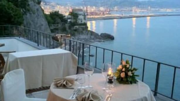 Un área de comedor al aire libre con mesas dispuestas para dos, con vistas a un paisaje urbano costero.