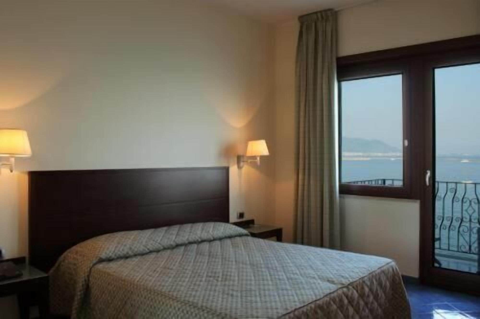 A hotel room with a large bed, two bedside lamps, a nightstand, a window with a view of the sea, and a balcony with a railing.