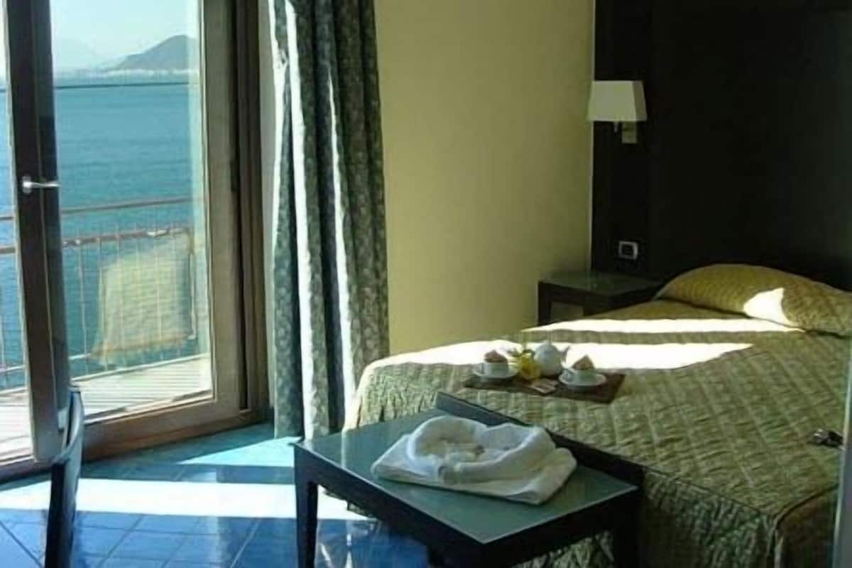 A hotel room with a bed, a table with a towel, a nightstand, a lamp, and a view of the sea through a window.