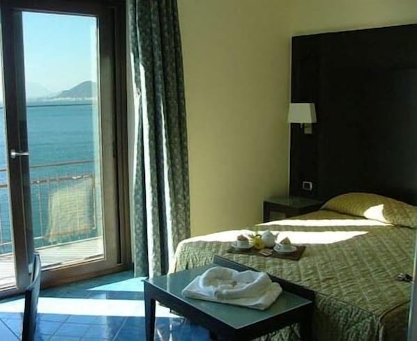 Habitación de hotel con cama, una mesa con una toalla, mesita de noche, lámpara y vistas al mar a través de una ventana.