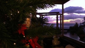 Un árbol de Navidad con luces y un lazo rojo, una ventana con vista al océano y al atardecer, y una mesita con un florero.