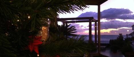 Un sapin de Noël décoré de guirlandes lumineuses et orné d’un nœud rouge, une fenêtre donnant sur l’océan et le coucher de soleil, et une petite table avec un vase.