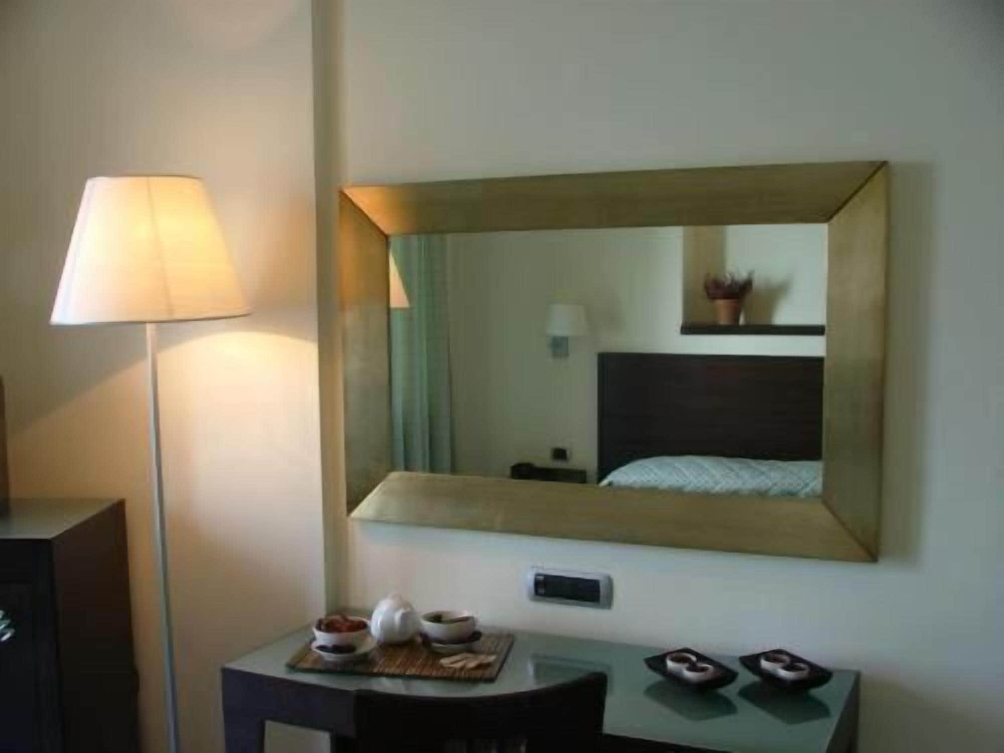 A hotel room with a bed, a bedside table with a lamp, a mirror, and a small table with a bowl and a teapot.