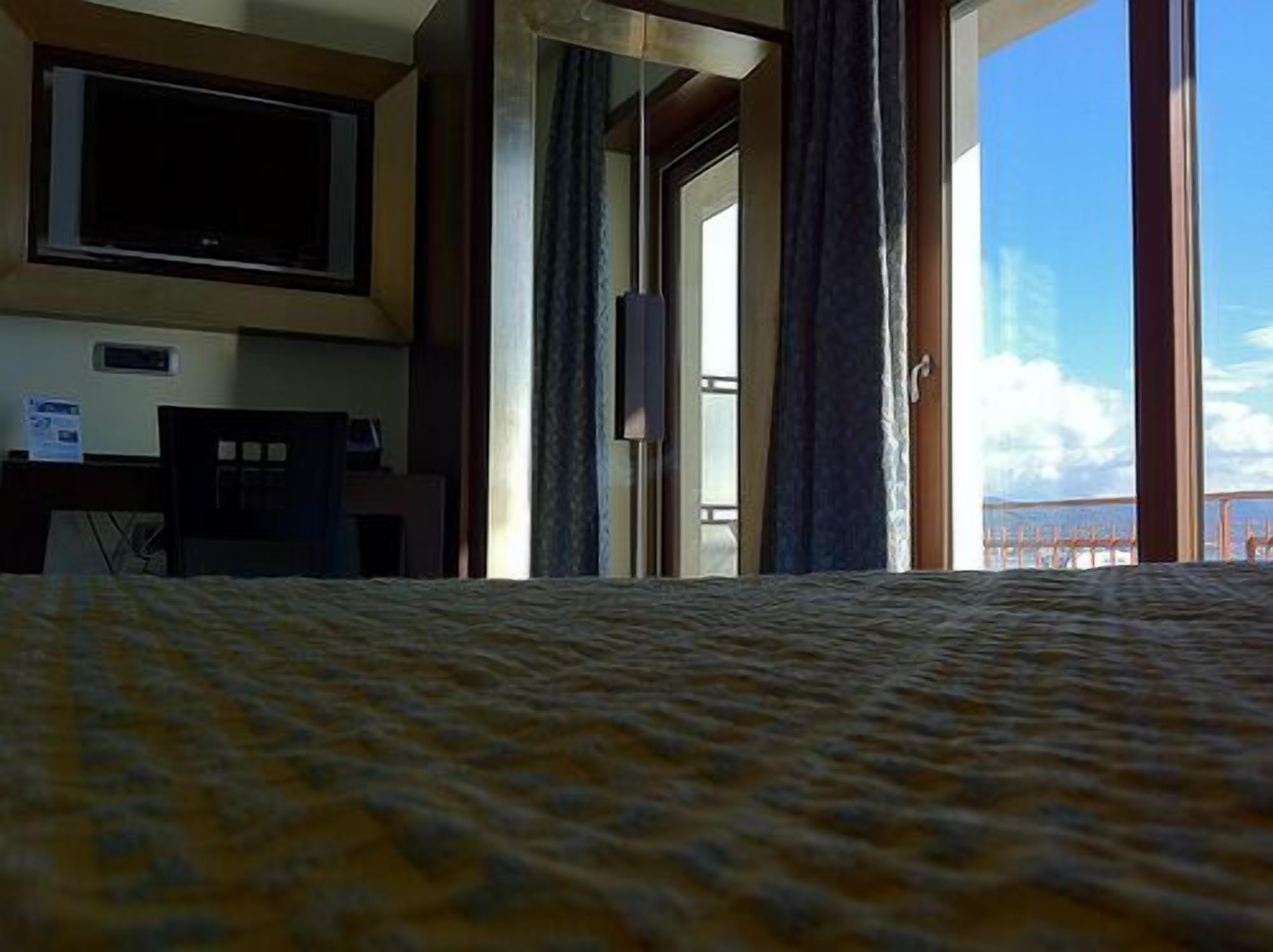 A hotel room with a bed, a television, a desk, and a view of the sea through large windows.